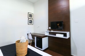 A modern hotel room with a wooden accent wall, a flat-screen TV mounted above a white shelf, a small desk with a chair, and a wicker basket on the floor. - Kak Garden Inn by Zuzu (Kuta)