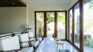 A modern dining area with a long white table, white cushioned seating, and large windows offering a view of the garden. - Kak Garden Inn by Zuzu (Kuta)