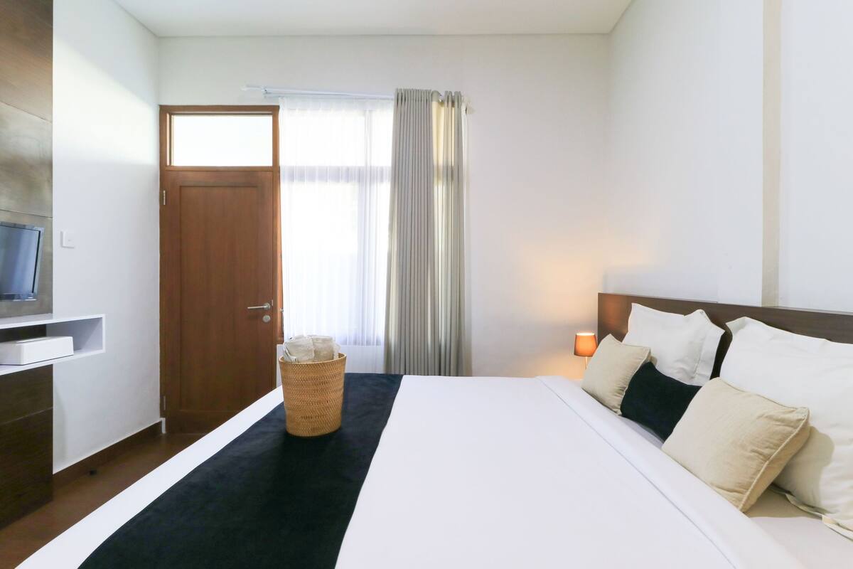 A hotel room with a bed, a TV, a wooden door, a window with curtains, and a nightstand with a lamp.