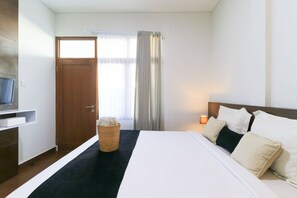 A hotel room with a bed, a TV, a wooden door, a window with curtains, and a nightstand with a lamp. - Kak Garden Inn by Zuzu (Kuta)