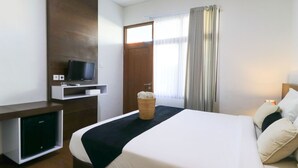 A hotel room with a bed, a TV on a stand, a small desk, and a window with curtains. - Kak Garden Inn by Zuzu (Kuta)