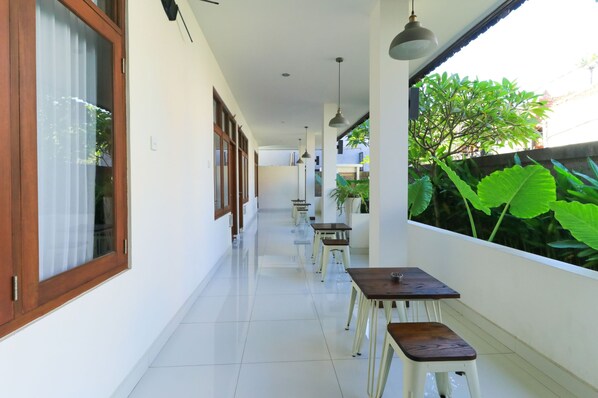 A corridor with tables and chairs, a view of greenery outside, and a window with a curtain. - Kak Garden Inn by Zuzu (Kuta)