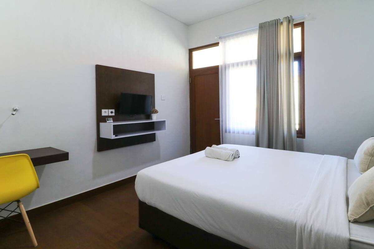 A hotel room with a bed, a TV mounted on the wall, a chair, and a window with curtains.