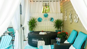Ett takförsett utomhusområde med sittplatser, utrustat med en uppblåsbar soffa från Coleman Saluspa, möbler i rotting och en dekorativ vägg med en skylt som visar ordet ”Beach”.