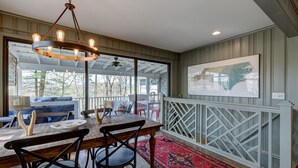 Un coin repas avec une table en bois, des chaises et un lustre. Une grande fenêtre offre une vue sur les arbres, et une terrasse se trouve à l’extérieur.