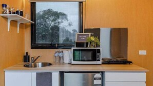 Eine kompakte Küche mit Spüle aus Edelstahl, einer Mikrowelle, einem kleinen Kühlschrank und einem Fenster mit Blick auf Bäume.