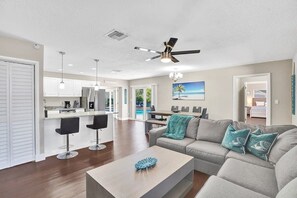 Ein modernes Wohnzimmer mit grauem Sofa, Couchtisch und Fernseher. Es gibt eine Küchenzeile mit weißen Schränken und einen Essbereich mit Blick auf einen Pool.