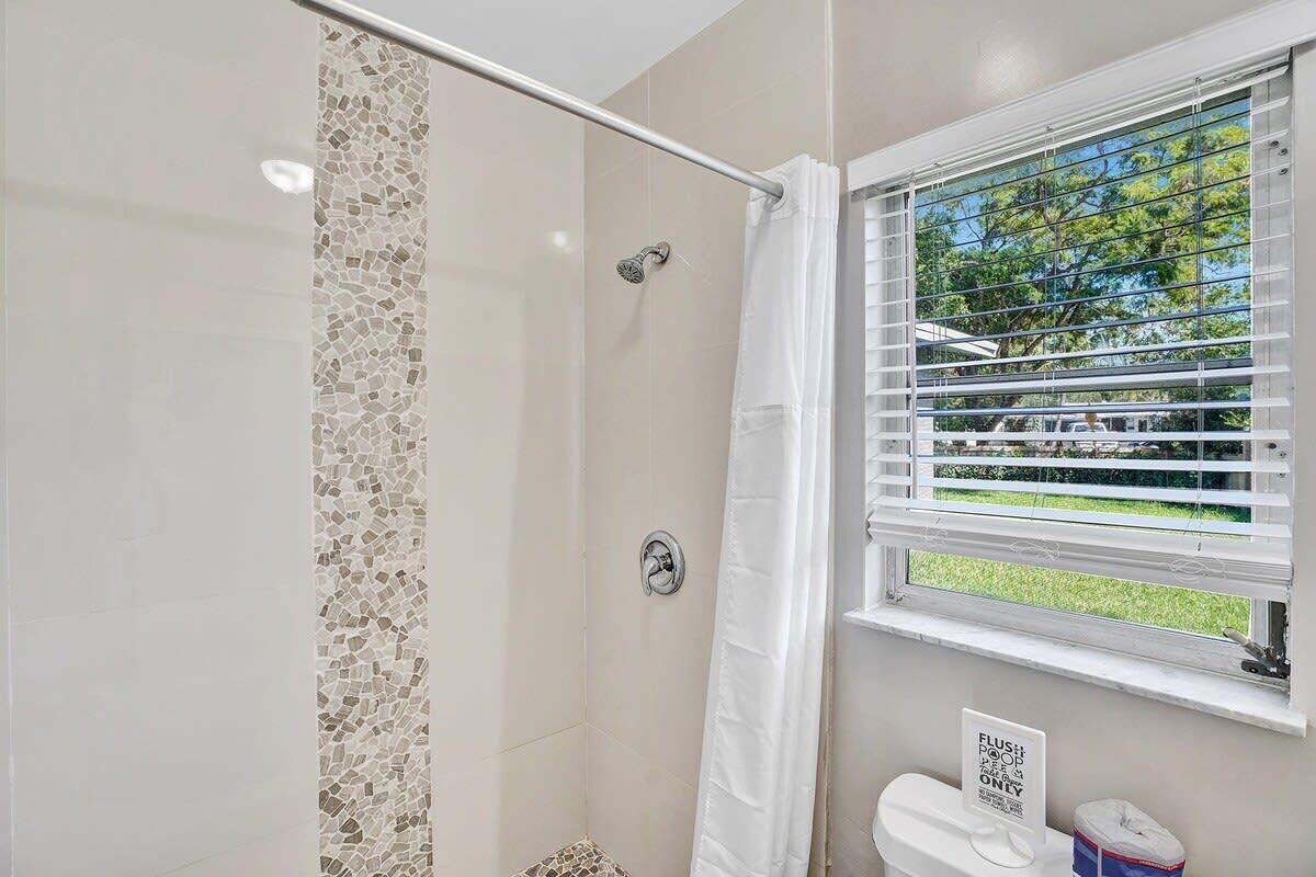 A bathroom with a shower, a toilet, a window with blinds, and a sign on the toilet.