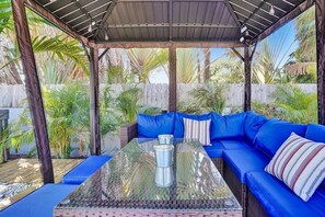 Un espace extérieur abrité, doté d’une table à plateau de verre, d’un canapé bleu à coussins moelleux et entouré de plantes tropicales.