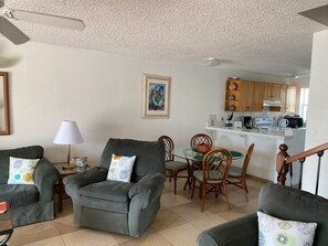 A living room with a sofa, armchairs, and a dining table with chairs. There is a kitchen in the background with wooden cabinets and a white countertop.