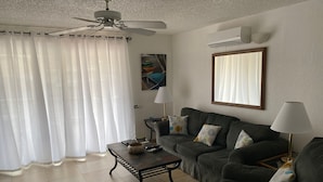 Una sala de estar con un sofá gris oscuro, una mesa de centro, un ventilador de techo y un ventanal con cortinas blancas.