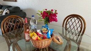Una mesa de comedor con cubierta de vidrio rodeada de sillas de madera, una cesta de mimbre con frutas y aperitivos, una botella de agua y un florero con flores.
