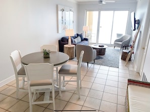 Uma sala de estar moderna com uma mesa de jantar, cadeiras, sofá e mesa de centro. Há uma janela grande que oferece luz natural e vista para o exterior.
