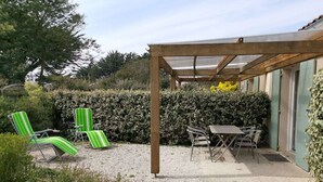 A covered patio area with a table and chairs, surrounded by a hedge and a gravel path. - Cottage Les Roses de mer ( Les Agapanthes ) (Sainte Marie de Ré)