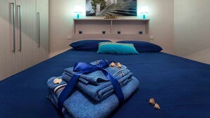 A neatly made bed with blue bedding, a blue pillow, and a folded towel with a blue ribbon. There are two wall-mounted lamps with white shades, and a framed picture of a building on the wall. - (MP63) Modern accommodation 150 mt from the sea (Marina Di Modica)