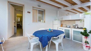 A compact kitchen with white cabinets, a stove, and a sink. There is a dining area with a table covered in a blue tablecloth and white chairs. A window with a view of the outside is visible, and a door leads to another room. - (MP63) Modern accommodation 150 mt from the sea (Marina Di Modica)