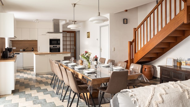 A modern kitchen with white cabinets, a dining area with a long table set for six, and a wooden staircase.