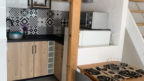 Een keuken met houten kastjes, een zwart aanrecht en tegelvloer met een patroon. Er staat een eettafel met stoelen en een hanglamp.