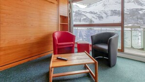 A room with a wooden wall, a red chair, a grey chair, a small table, and a view of snow-covered mountains through large windows.