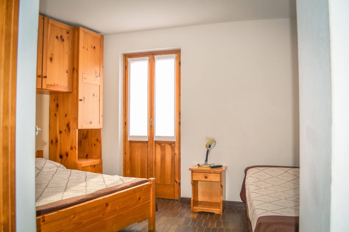 A bedroom with two wooden beds, a wooden wardrobe, a small bedside table with a lamp, and a window with blinds.