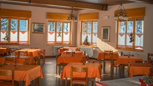 A dining area with multiple tables and chairs, large windows with curtains, and a wooden ceiling. - Hotel Milano (Valfurva)