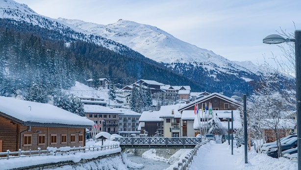 A snow-covered village with wooden buildings, a bridge, and a mountain backdrop. - Hotel Milano (Valfurva)