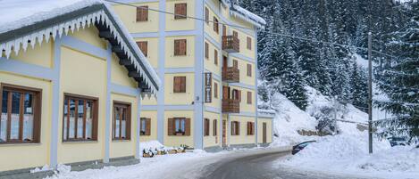 Un edificio de varios pisos con fachada amarilla y contraventanas de madera, rodeado de árboles cubiertos de nieve y un paisaje nevado.