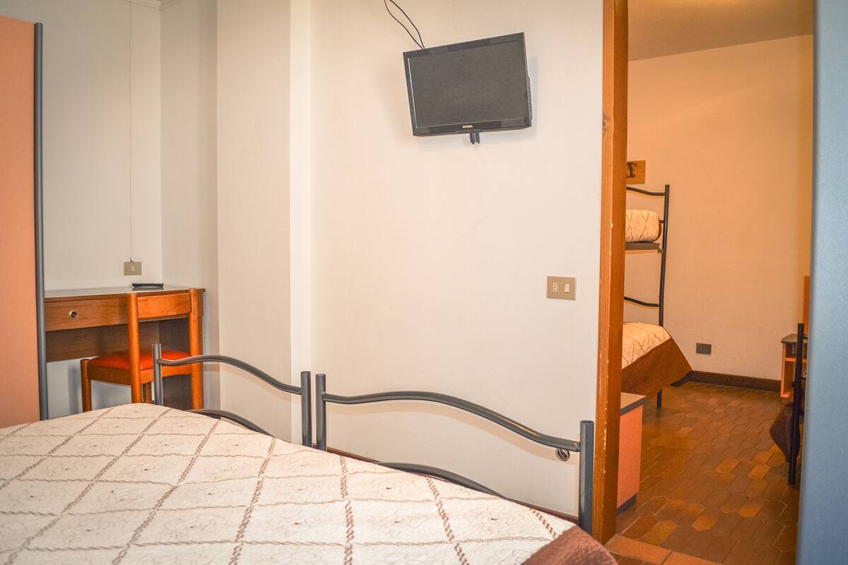 A bedroom with a bed, a desk, a chair, a television mounted on the wall, and a bunk bed in the adjacent room.