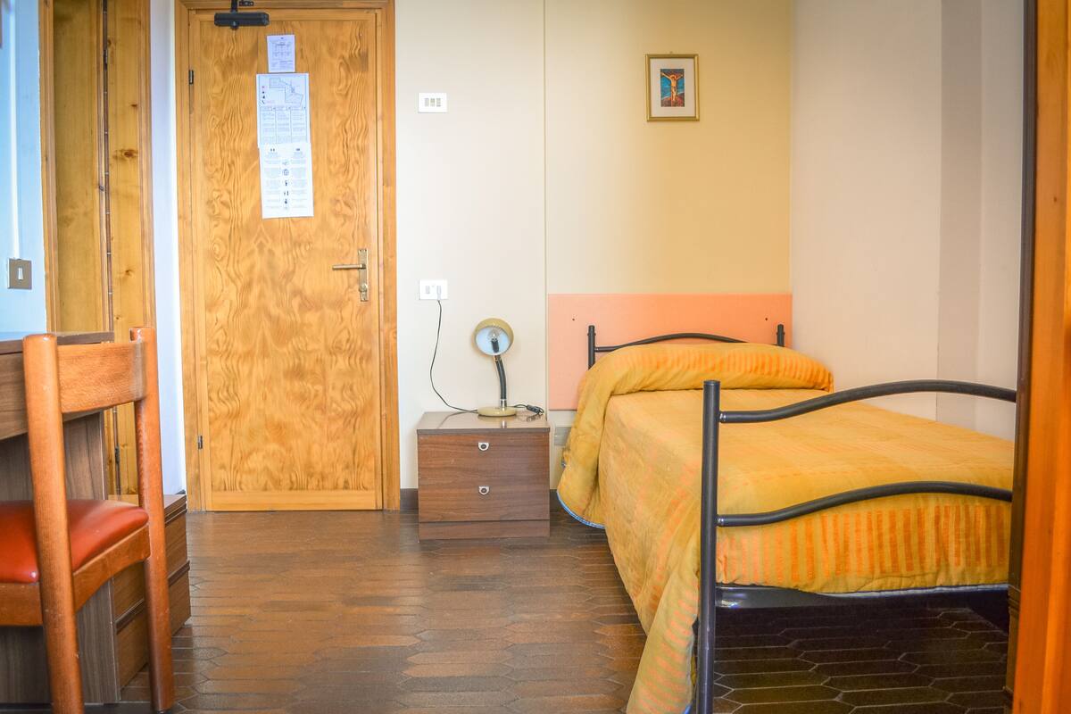 A bedroom with a wooden door, a bedside table with a lamp, a bed with an orange bedspread, and a chair.