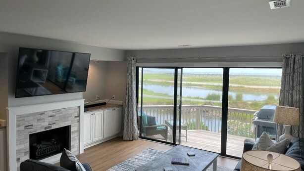 En moderne stue med pejs, et fladskærms-tv og en stor skydedør, der fører ud til en terrasse med udsigt over en sø.