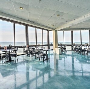 A spacious dining area with large windows overlooking the sea, featuring tables and chairs arranged for dining.