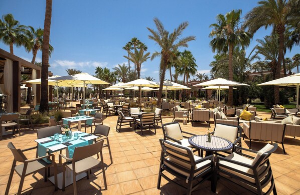 An outdoor patio area with tables and chairs, palm trees, and umbrellas. - Sol Oasis Marrakech - All Inclusive (Marrakech)