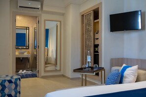 A hotel room with a bed, a TV mounted on the wall, a bedside table with glasses and a bottle, a mirror, and a wardrobe.