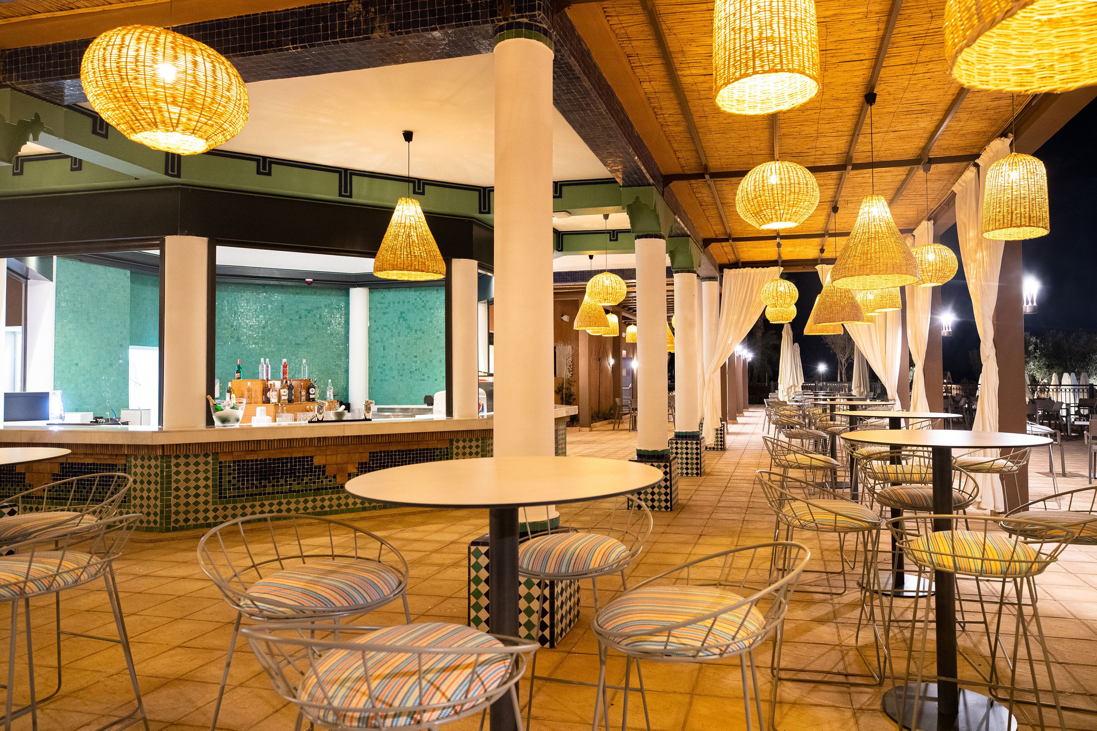 An outdoor dining area with round tables and metal chairs, surrounded by white pillars and hanging wicker lamps.