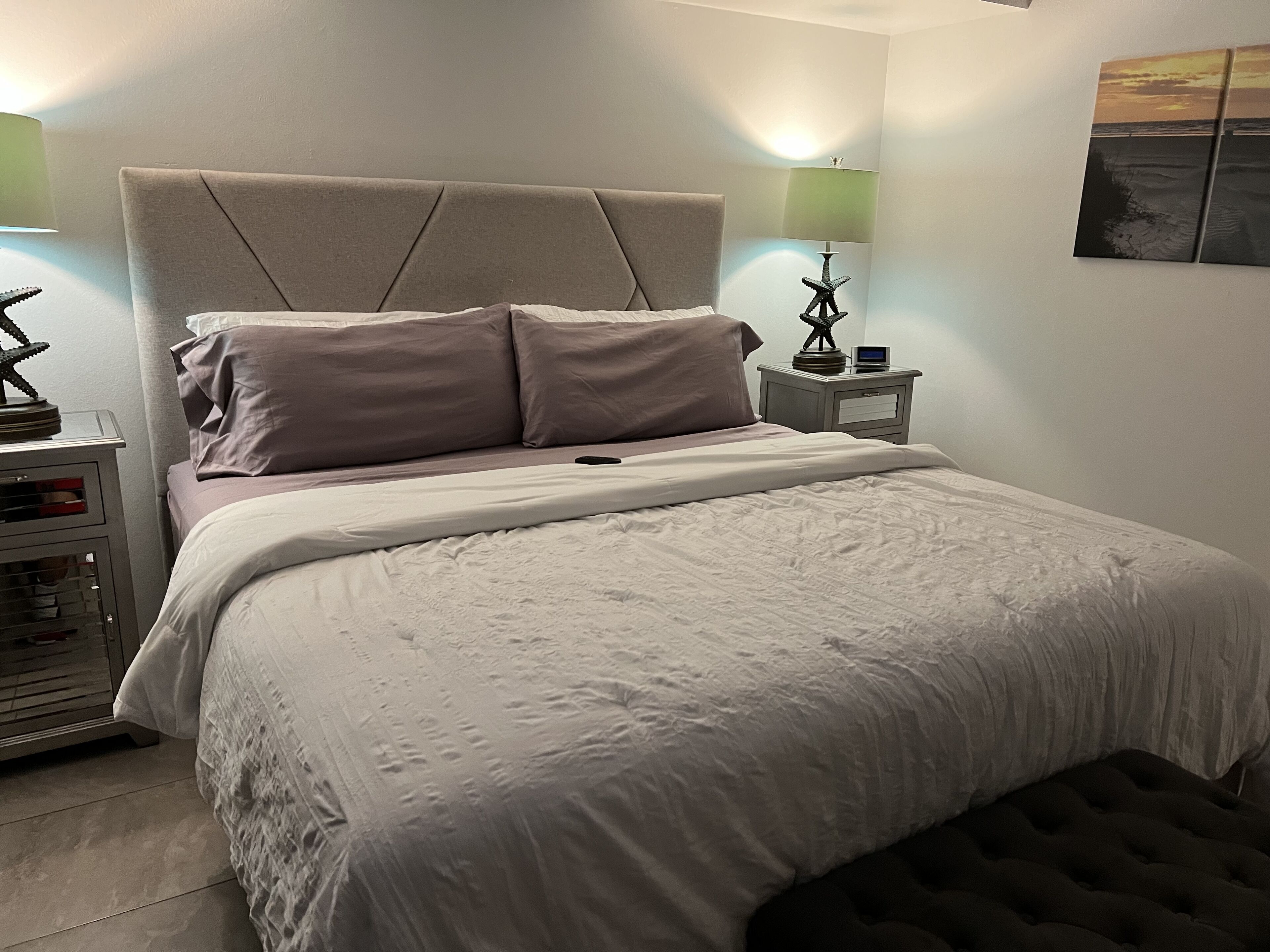 Ein ordentlich bezogenes Bett mit Kopfteil, zwei Nachttischlampen, einem kleinen Tisch mit einem dekorativen Gegenstand und einem Wandbild.