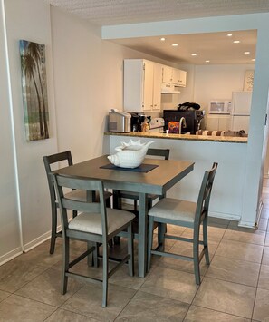 Uma sala de jantar com mesa e cadeiras, uma cozinha ao fundo, e um quadro na parede.