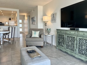 Uma sala de estar com uma poltrona cinza, uma mesinha com abajur, uma TV e um armário com detalhes elaborados.