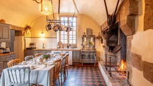 A traditional kitchen with a stone fireplace, a dining table set for a meal, and a checkered floor. - Château des Louteaux (Chézy)