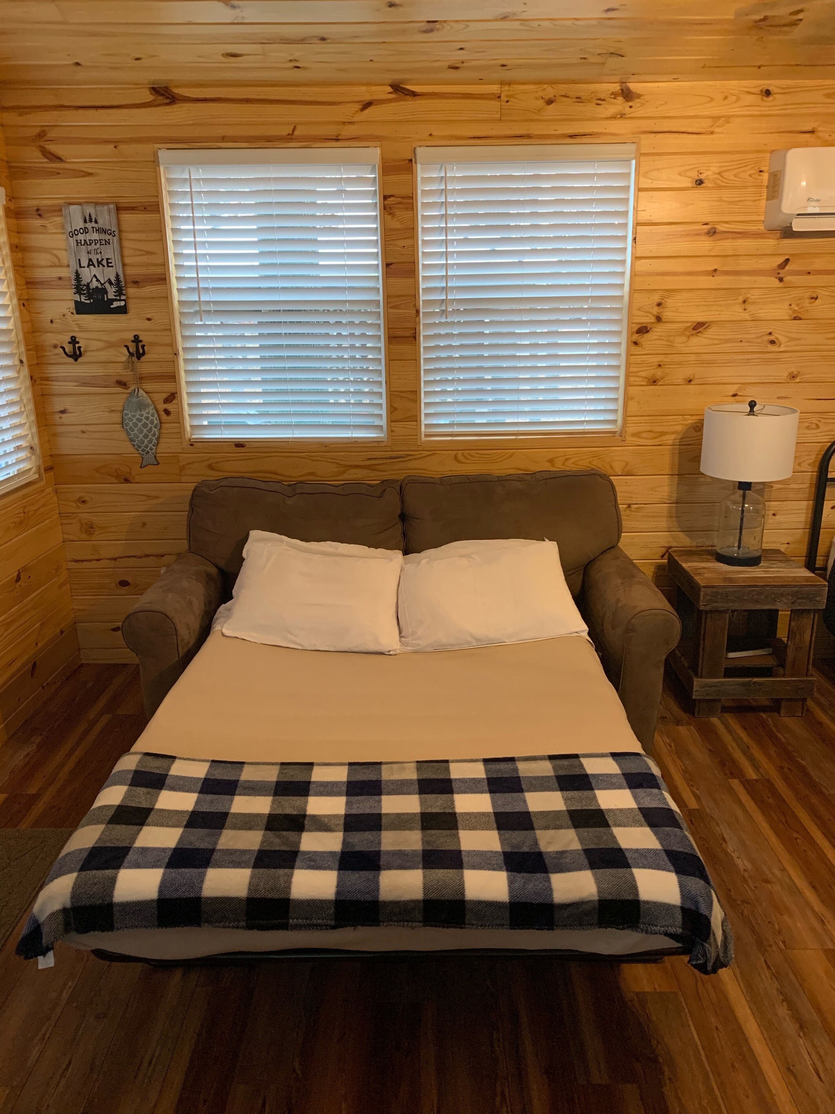 A cozy log cabin bedroom with a bed, two windows with blinds, a lamp on a side table, and a checkered throw blanket.