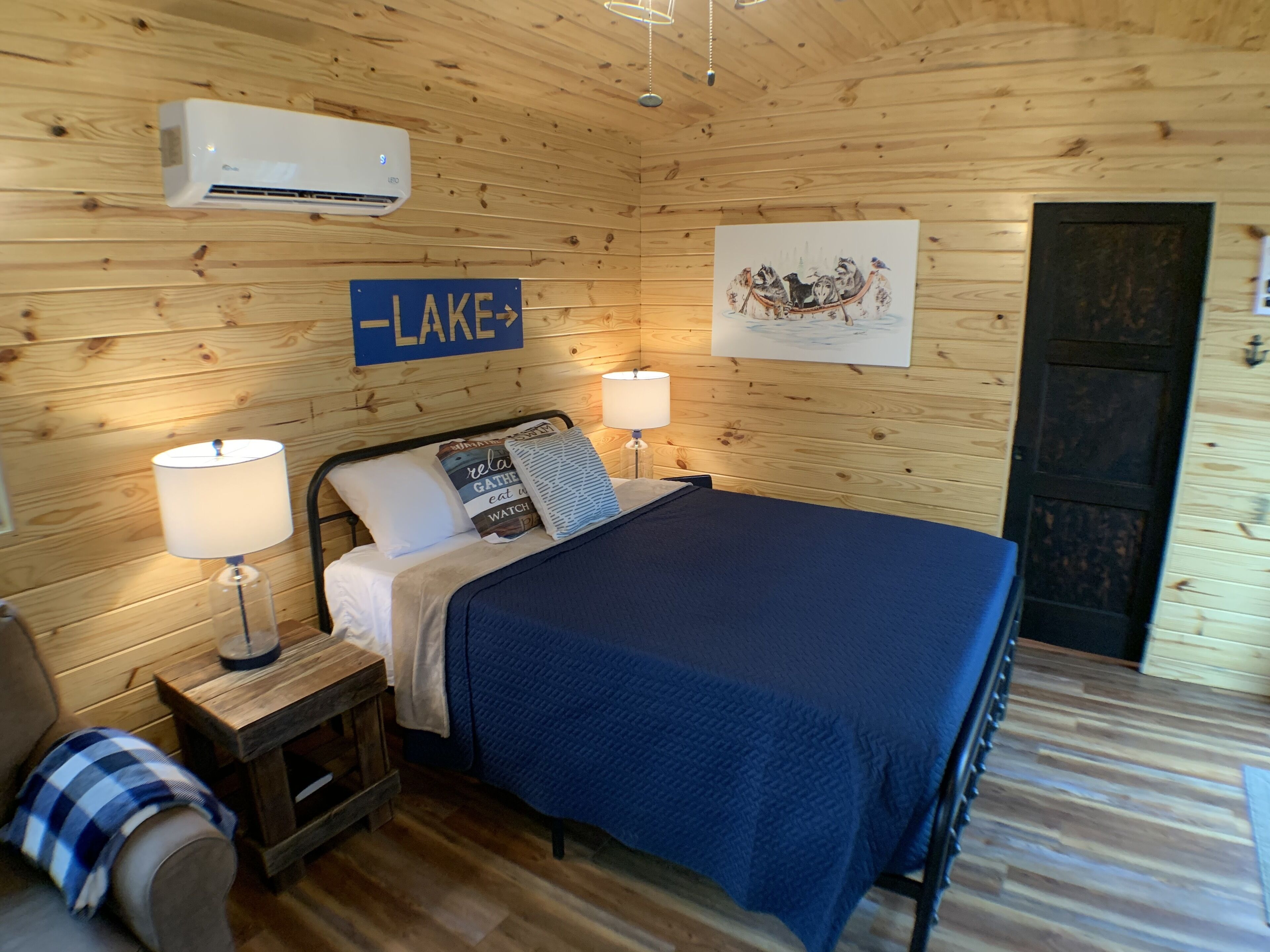 A wooden-paneled room with a bed, two bedside lamps, a sign reading "LAKE", and a painting of a boat on the wall.
