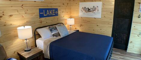 A wooden-paneled room with a bed, two bedside lamps, a sign reading "LAKE", and a painting of a boat on the wall.