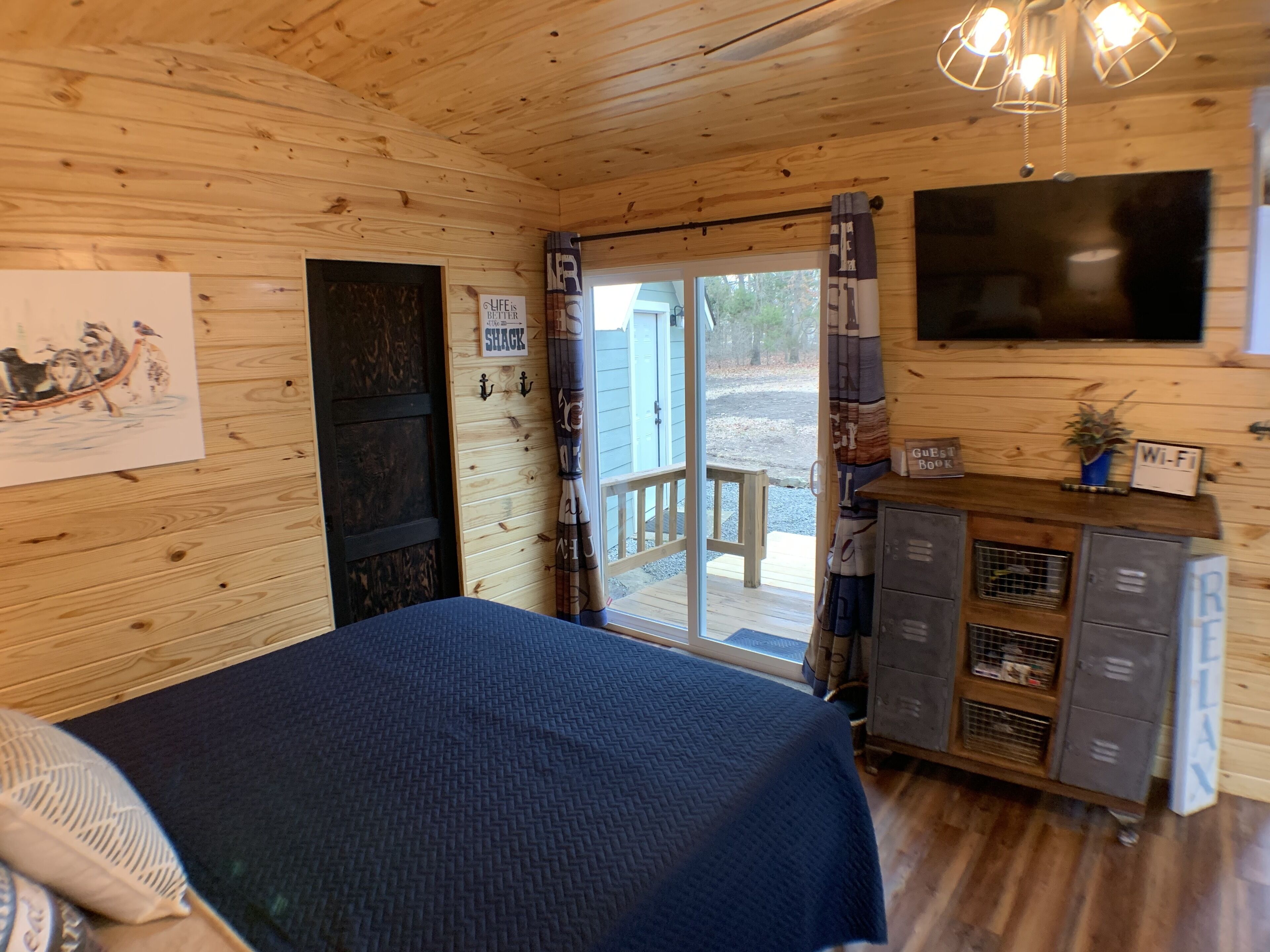 Habitación de cabaña de madera con una cama, un televisor, un escritorio con una computadora y una puerta que da al exterior.