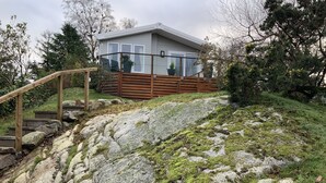 A house with a wooden staircase leading up to a deck, surrounded by large rocks and greenery. - Clouded Yellow - Beautiful Modern Lodge, Kippford (Dalbeattie)