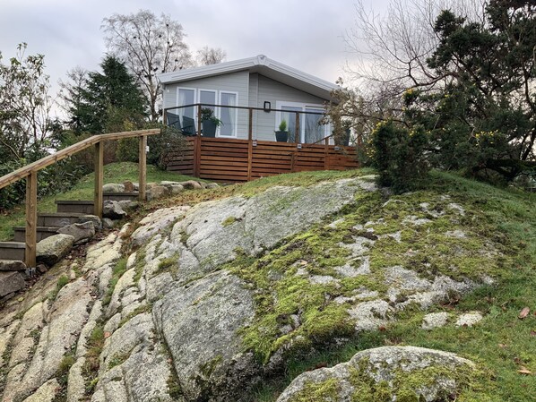 A house with a wooden staircase leading up to a deck, surrounded by large rocks and greenery.