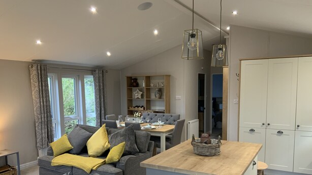A modern living room with a grey sofa, yellow cushions, a wooden dining table, and a kitchen area with white cabinets. - Clouded Yellow - Beautiful Modern Lodge, Kippford (Dalbeattie)