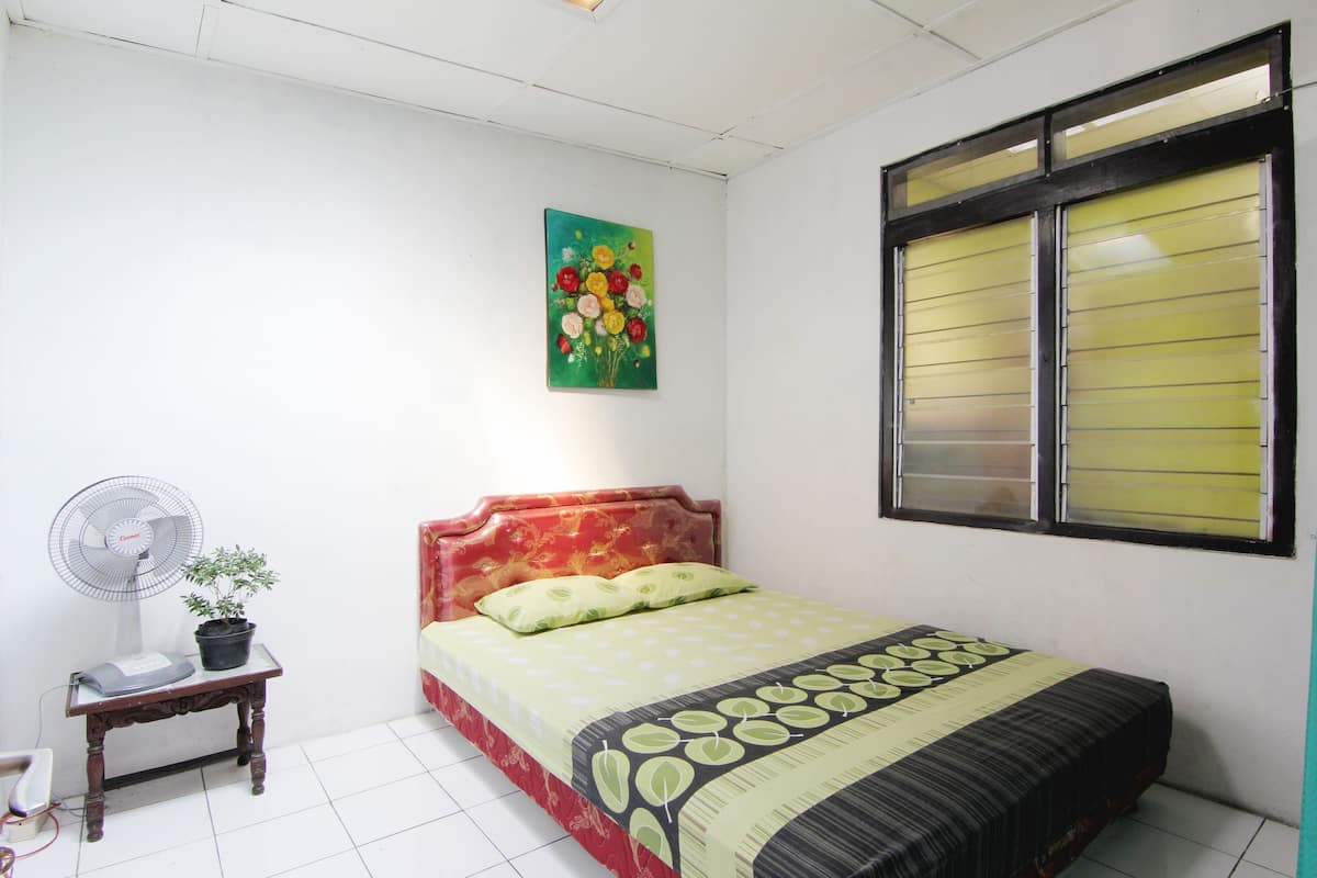 A bedroom with a bed, a bedside table with a fan and a plant, a window with blinds, and a painting on the wall.