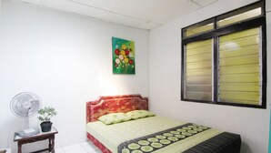 Un dormitorio con una cama, una mesita de noche con un ventilador y una planta, una ventana con persianas y un cuadro en la pared.