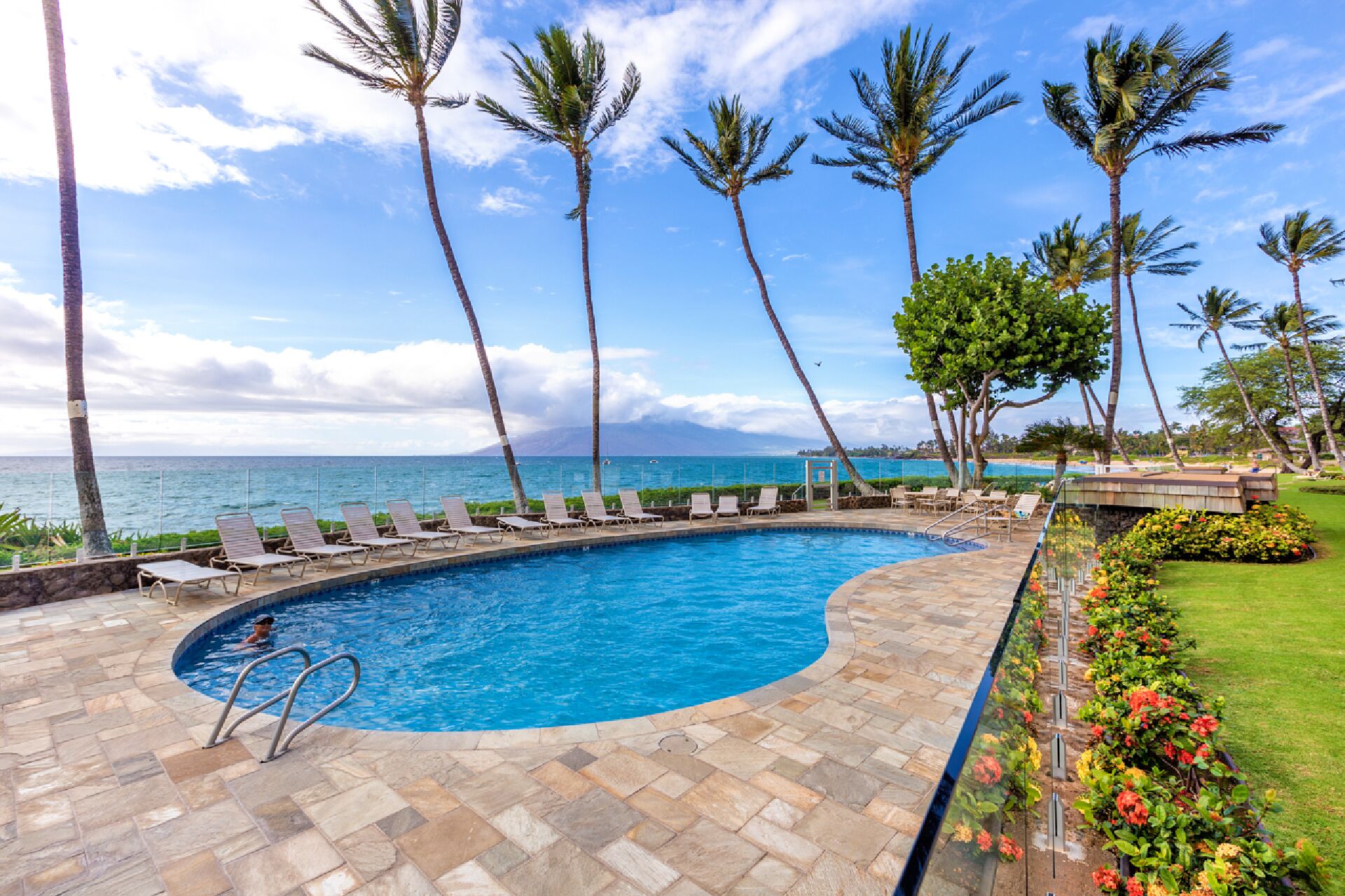 A pool area with lounge chairs, palm trees, and a view of the ocean.