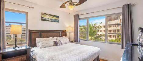 A hotel room with a large bed, two bedside tables with lamps, a ceiling fan, and large windows offering a view of palm trees and buildings.