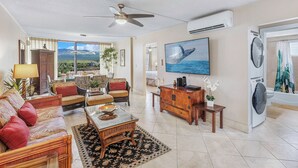 A spacious living room with a sofa, chairs, a coffee table, and a television. There is a large window with a view of mountains and a ceiling fan. - Remodeled Luxury at Royal Mauian | Haleakala Views and Oceanfront Living (Kihei)